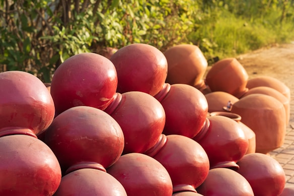 A collection of red clay pots stacked neatly outdoors with a natural setting in the background. The pots have a glossy finish, and the sunlight creates a warm glow on their surfaces. Green foliage and a cobblestone path are visible in the background.