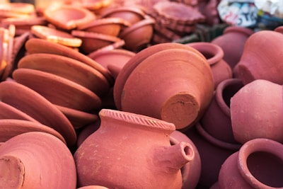 A collection of reddish-brown clay pots lies in a heap, displaying a variety of shapes including bowls and pitchers. The pots have natural textures and earthy tones, with some showing intricate designs around their rims.