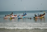 Colorful fishing boats are anchored in shallow water, with several people working and swimming around them. The ocean is calm, and a few birds are flying overhead. The horizon shows distant ships, and the scene captures an activity-rich coastal environment.