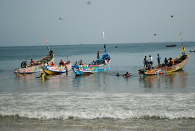 Colorful fishing boats are anchored in shallow water, with several people working and swimming around them. The ocean is calm, and a few birds are flying overhead. The horizon shows distant ships, and the scene captures an activity-rich coastal environment.