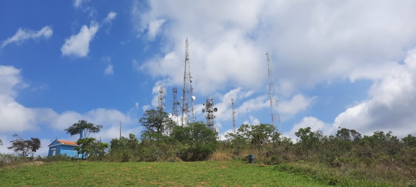 Telecommunication towers surrounded by lush greenery with a clear blue sky and scattered clouds above. A small blue building with a red roof is visible on the left side, partly obscured by trees.