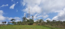 Telecommunication towers surrounded by lush greenery with a clear blue sky and scattered clouds above. A small blue building with a red roof is visible on the left side, partly obscured by trees.