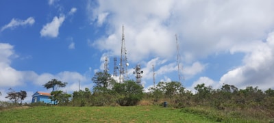 Telecommunication towers surrounded by lush greenery with a clear blue sky and scattered clouds above. A small blue building with a red roof is visible on the left side, partly obscured by trees.