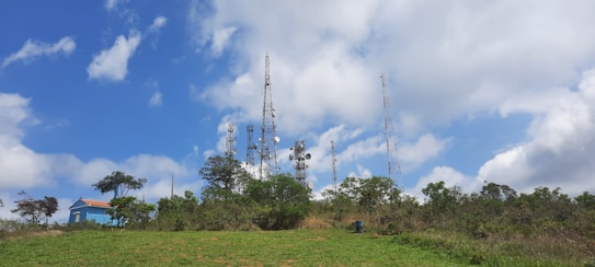 Telecommunication towers surrounded by lush greenery with a clear blue sky and scattered clouds above. A small blue building with a red roof is visible on the left side, partly obscured by trees.