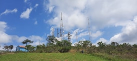 Telecommunication towers surrounded by lush greenery with a clear blue sky and scattered clouds above. A small blue building with a red roof is visible on the left side, partly obscured by trees.