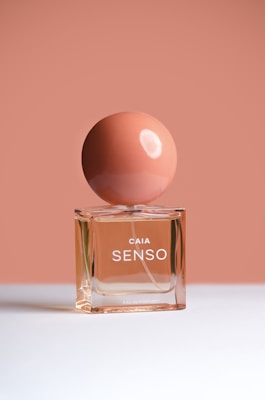 A perfume bottle with a round cap is placed against a minimalist background. The container is transparent, showcasing a peach-colored liquid, while the matte spherical cap is a slightly darker shade of peach. The bottle is labeled 'CAIA SENSO' in white letters.