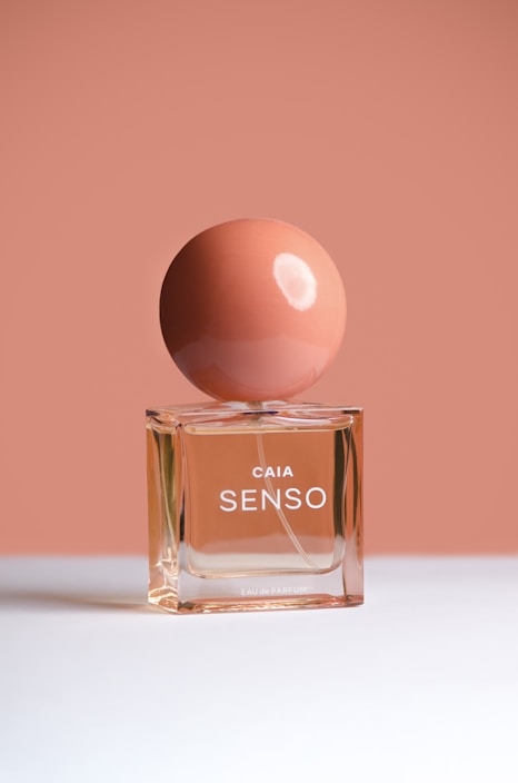 A perfume bottle with a round cap is placed against a minimalist background. The container is transparent, showcasing a peach-colored liquid, while the matte spherical cap is a slightly darker shade of peach. The bottle is labeled 'CAIA SENSO' in white letters.