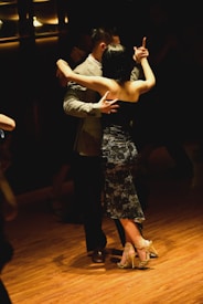 A couple elegantly dancing in a dimly lit room, with the woman wearing a black lace dress and high heels. The man is dressed in a light-colored suit. The atmosphere conveys an intimate and graceful mood.