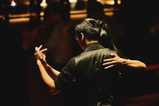 A couple is captured in a close embrace while dancing in a dimly lit setting. The focus is on the dancers' hands and upper bodies, highlighting the intimacy and grace of the moment. The background is softly blurred, suggesting a cozy and atmospheric environment, perhaps in a dance hall or club.