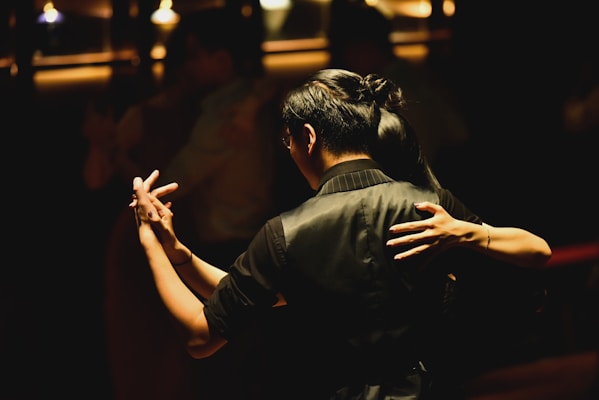 A couple is captured in a close embrace while dancing in a dimly lit setting. The focus is on the dancers' hands and upper bodies, highlighting the intimacy and grace of the moment. The background is softly blurred, suggesting a cozy and atmospheric environment, perhaps in a dance hall or club.