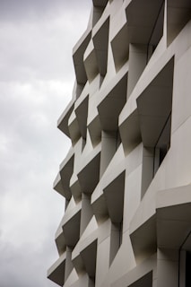 The image features an abstract architectural design with repeating geometric patterns on the facade of a building. The structure consists of protruding rectangular and trapezoidal elements casting distinct shadows, creating a three-dimensional effect.