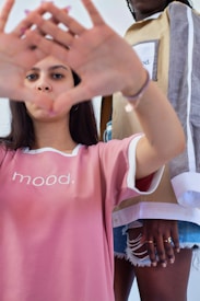 Two individuals are portrayed in casual, trendy fashion. The person in the foreground is wearing a pink shirt with the word 'mood' printed on it and is making a hand gesture forming a triangle shape. The second individual, partially visible, is wearing a stylish outfit featuring layered clothing with a similar 'mood' print.