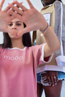 Two individuals are portrayed in casual, trendy fashion. The person in the foreground is wearing a pink shirt with the word 'mood' printed on it and is making a hand gesture forming a triangle shape. The second individual, partially visible, is wearing a stylish outfit featuring layered clothing with a similar 'mood' print.