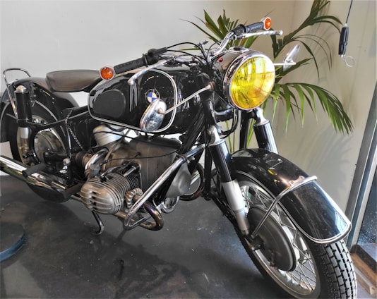 A vintage motorcycle with a shiny black finish and chrome accents is displayed indoors. The motorcycle features a prominent yellow-tinted headlight and exposed engine components. The handlebars are equipped with rearview mirrors, and the tires exhibit a classic tread pattern. In the background, green foliage adds a touch of nature to the setting, which appears well-lit by natural light coming through a nearby window.