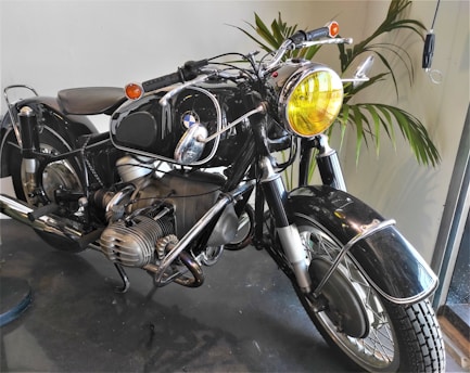 A vintage motorcycle with a shiny black finish and chrome accents is displayed indoors. The motorcycle features a prominent yellow-tinted headlight and exposed engine components. The handlebars are equipped with rearview mirrors, and the tires exhibit a classic tread pattern. In the background, green foliage adds a touch of nature to the setting, which appears well-lit by natural light coming through a nearby window.