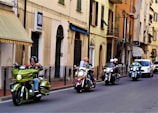 A group of motorcyclists ride down a street lined with Italian-style buildings. The road is flanked by various shops and residences, with visible signs and balconies. The bikers wear helmets and some have windshields, suggesting a casual city ride.