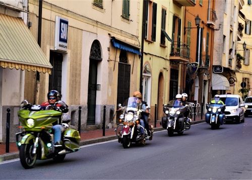 A group of motorcyclists ride down a street lined with Italian-style buildings. The road is flanked by various shops and residences, with visible signs and balconies. The bikers wear helmets and some have windshields, suggesting a casual city ride.