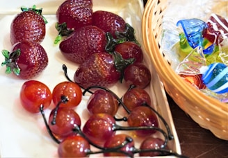 Glass figurines shaped like fruits, specifically strawberries and cherries, are artistically arranged on a light-colored surface. To the right, a wicker basket contains an assortment of colorful, decorative glass pieces resembling wrapped candies.