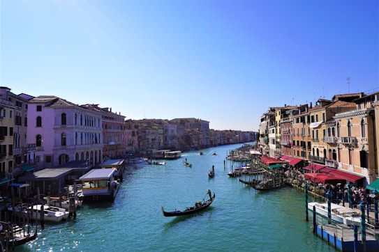 A picturesque canal flanked by historic buildings with colorful facades and red awnings on both sides. Several gondolas and small boats navigate the calm, turquoise waters under a clear blue sky.