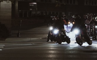 A group of motorcyclists is riding their bikes at night, illuminated by the glare of headlights. The scene appears dynamic, with the sense of motion and the smoke trailing behind the bikes. Some riders wear bunny suits, adding an unusual and playful element to the scene.
