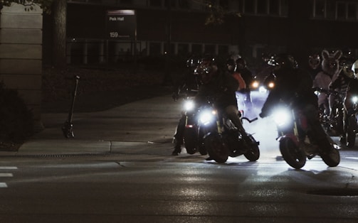 A group of motorcyclists is riding their bikes at night, illuminated by the glare of headlights. The scene appears dynamic, with the sense of motion and the smoke trailing behind the bikes. Some riders wear bunny suits, adding an unusual and playful element to the scene.