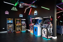 An arcade room with a row of classic arcade game machines including titles like 'Mortal Kombat' and 'RoboCop'. Vibrant neon lights and graphics of iconic 80s video game characters such as Pac-Man and Space Invaders illuminate the dark room. A large duct runs across the ceiling, adding an industrial feel.