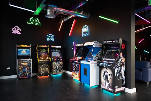 An arcade room with a row of classic arcade game machines including titles like 'Mortal Kombat' and 'RoboCop'. Vibrant neon lights and graphics of iconic 80s video game characters such as Pac-Man and Space Invaders illuminate the dark room. A large duct runs across the ceiling, adding an industrial feel.