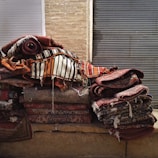 A stack of colorful, woven rugs with intricate patterns is piled against a backdrop of brick and metal shutters. The rugs display various designs, from stripes to floral motifs, with a predominantly warm color palette.