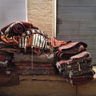 A stack of colorful, woven rugs with intricate patterns is piled against a backdrop of brick and metal shutters. The rugs display various designs, from stripes to floral motifs, with a predominantly warm color palette.