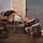 A stack of colorful, woven rugs with intricate patterns is piled against a backdrop of brick and metal shutters. The rugs display various designs, from stripes to floral motifs, with a predominantly warm color palette.
