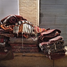 A stack of colorful, woven rugs with intricate patterns is piled against a backdrop of brick and metal shutters. The rugs display various designs, from stripes to floral motifs, with a predominantly warm color palette.