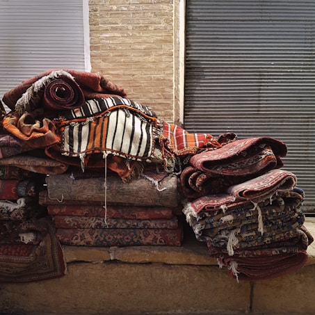 A stack of colorful, woven rugs with intricate patterns is piled against a backdrop of brick and metal shutters. The rugs display various designs, from stripes to floral motifs, with a predominantly warm color palette.