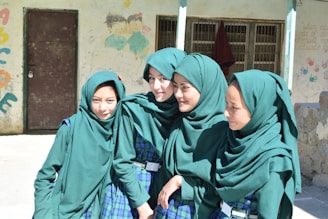 Four young girls stand closely together, smiling and wearing matching green headscarves and plaid uniforms with green accents. They are in an outdoor setting, with a wall behind them that features colorful graffiti and children's drawings. A brown door and barred window are visible in the background.