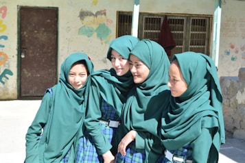 Four young girls stand closely together, smiling and wearing matching green headscarves and plaid uniforms with green accents. They are in an outdoor setting, with a wall behind them that features colorful graffiti and children's drawings. A brown door and barred window are visible in the background.