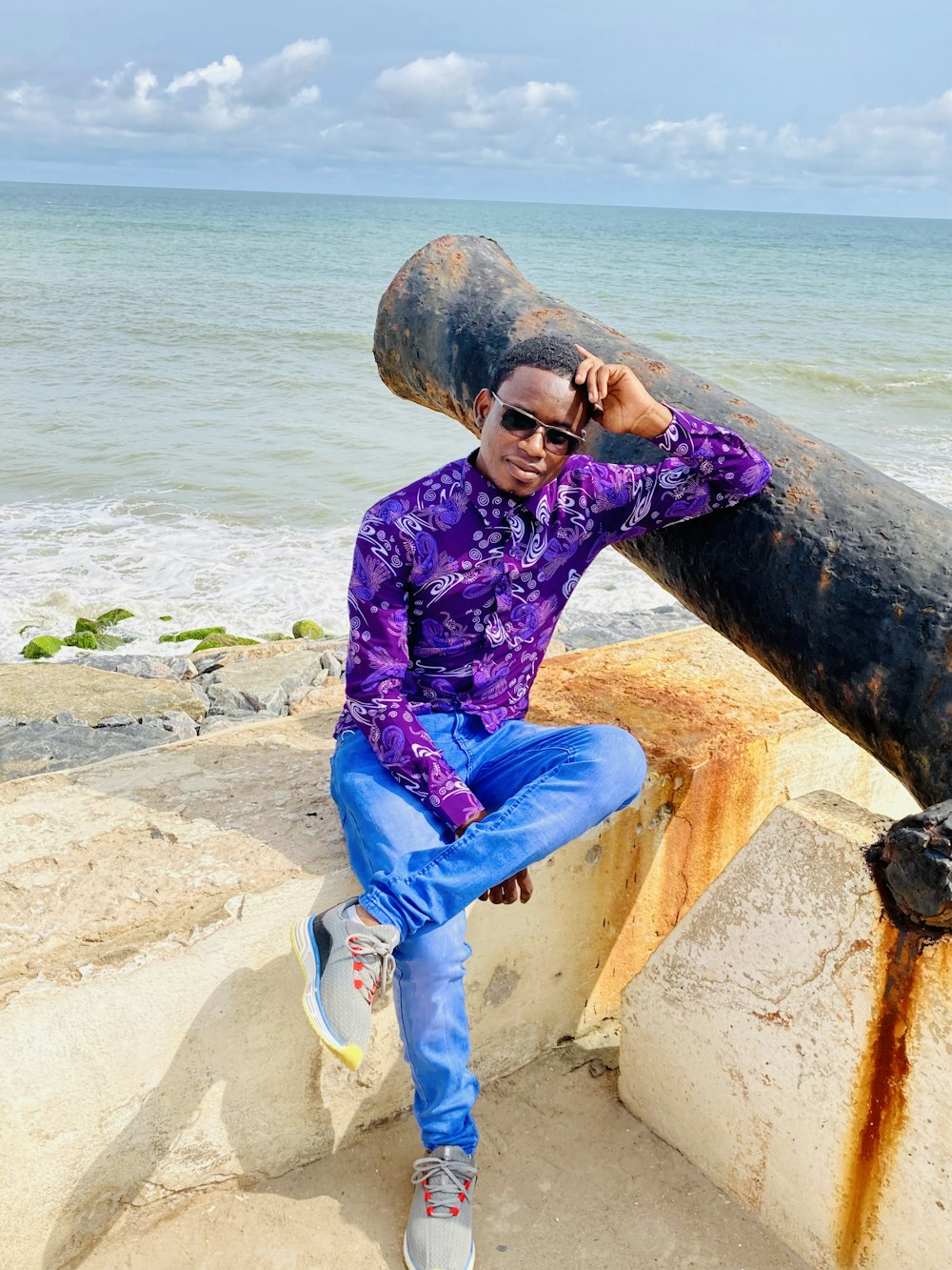 a man in a purple shirt and blue pants sitting on a rock near the ocean