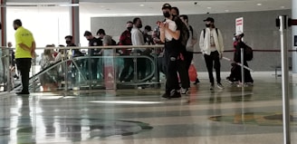 A group of people stands near an escalator in a large indoor space. Most are wearing face masks and casual clothing, with one person in a bright yellow 'Security' shirt observing from the side. The area has polished, patterned flooring and overhead lighting.