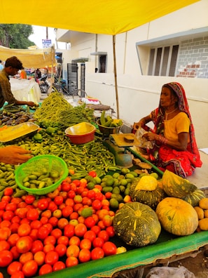 A vibrant vegetable stall at an outdoor market features an assortment of fresh produce including red tomatoes, green beans, cucumbers, and large pumpkins. A woman in colorful traditional attire is sitting behind the stall, attending to customers under a bright yellow canopy.