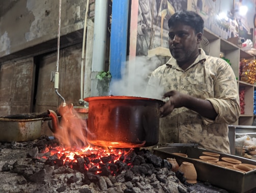 A man is preparing a hot beverage using a large pot over an open coal fire. The fire is producing bright red embers, and steam rises from the pot. Several small clay pots are placed nearby. The setting appears to be rustic, with a simple kitchen setup and some shelves in the background.