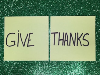 Two yellow post-it notes placed side by side on a glittery green background. The left note has the word 'GIVE' written on it and the right note has the word 'THANKS'.