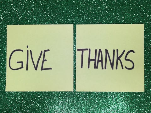 Two yellow post-it notes placed side by side on a glittery green background. The left note has the word 'GIVE' written on it and the right note has the word 'THANKS'.