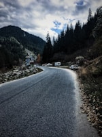 A winding road leads through a mountainous landscape with dense evergreen forests lining the sides. A few vehicles, including a campervan, are parked near a small structure resembling a castle or old building. Cloudy skies loom overhead, creating a dramatic and mysterious atmosphere.