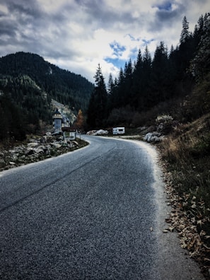 A winding road leads through a mountainous landscape with dense evergreen forests lining the sides. A few vehicles, including a campervan, are parked near a small structure resembling a castle or old building. Cloudy skies loom overhead, creating a dramatic and mysterious atmosphere.