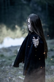 A person wearing a black cloak presents a mysterious and intense pose, with long hair partially covering their face. The background is an outdoor setting, somewhat blurred, lending a sense of intrigue and focus on the figure.