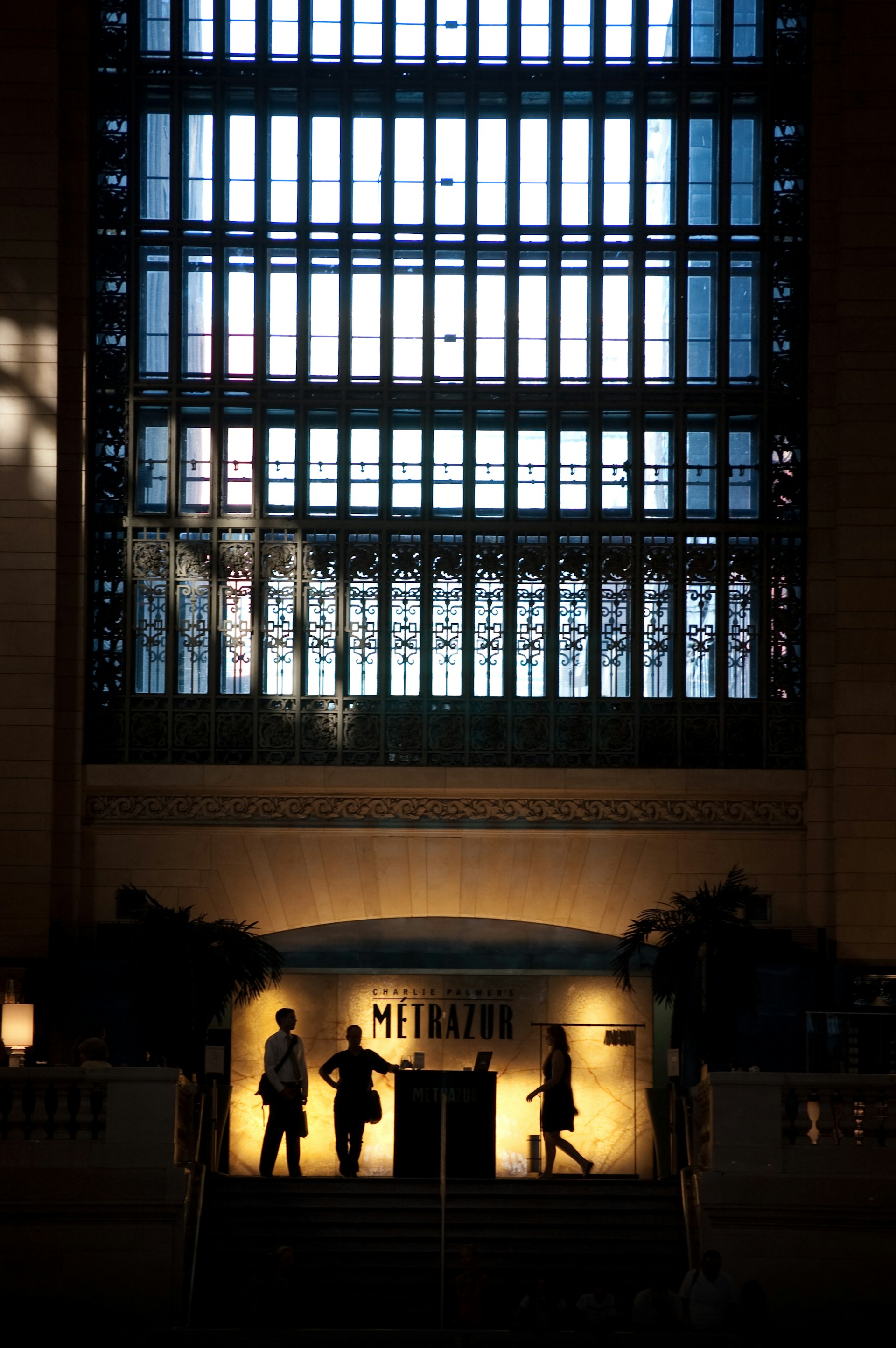 A grand interior space featuring a large, ornate window with intricate metalwork and a high ceiling. Silhouettes of three people are seen walking past a brightly lit archway with the word 'Metrazur' prominently displayed. The atmosphere is sophisticated and classic, with a mix of historical and modern elements.
