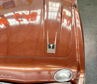 A vintage car hood with a deep brown, metallic finish featuring reflective highlights. The hood displays a sleek emblem with a stylized 'S' and twin chrome lines. Visible through the reflection are neon red letters and the industrial ceiling of a garage or showroom.