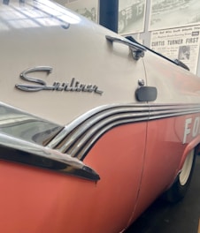 A close-up view of a vintage car with a pink and white color scheme. The car features chrome detailing and the word 'Sunliner' emblazoned on the side. The background includes a display of vintage racing posters and newspaper articles.