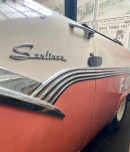 A close-up view of a vintage car with a pink and white color scheme. The car features chrome detailing and the word 'Sunliner' emblazoned on the side. The background includes a display of vintage racing posters and newspaper articles.