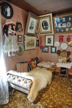 A cozy interior space featuring a vintage-style daybed covered with a white fur throw and colorful decorative pillows. The surrounding walls are adorned with an eclectic mix of framed photographs and paintings in various sizes, including portraits and vintage images. A large, ornate carpet covers the floor, and a small wooden table topped with lace fabric displays small decorative items.