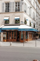 A charming corner café with blue awnings and large glass windows located in a European-style building. The structure features light-colored stonework and traditional shutters, with neatly trimmed bushes decorating the ledges. The street in front is quiet with minimal pedestrian or vehicular traffic.