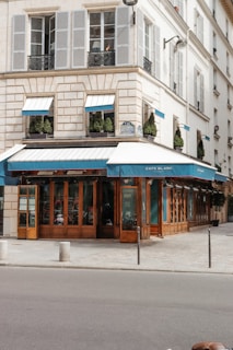 A charming corner café with blue awnings and large glass windows located in a European-style building. The structure features light-colored stonework and traditional shutters, with neatly trimmed bushes decorating the ledges. The street in front is quiet with minimal pedestrian or vehicular traffic.